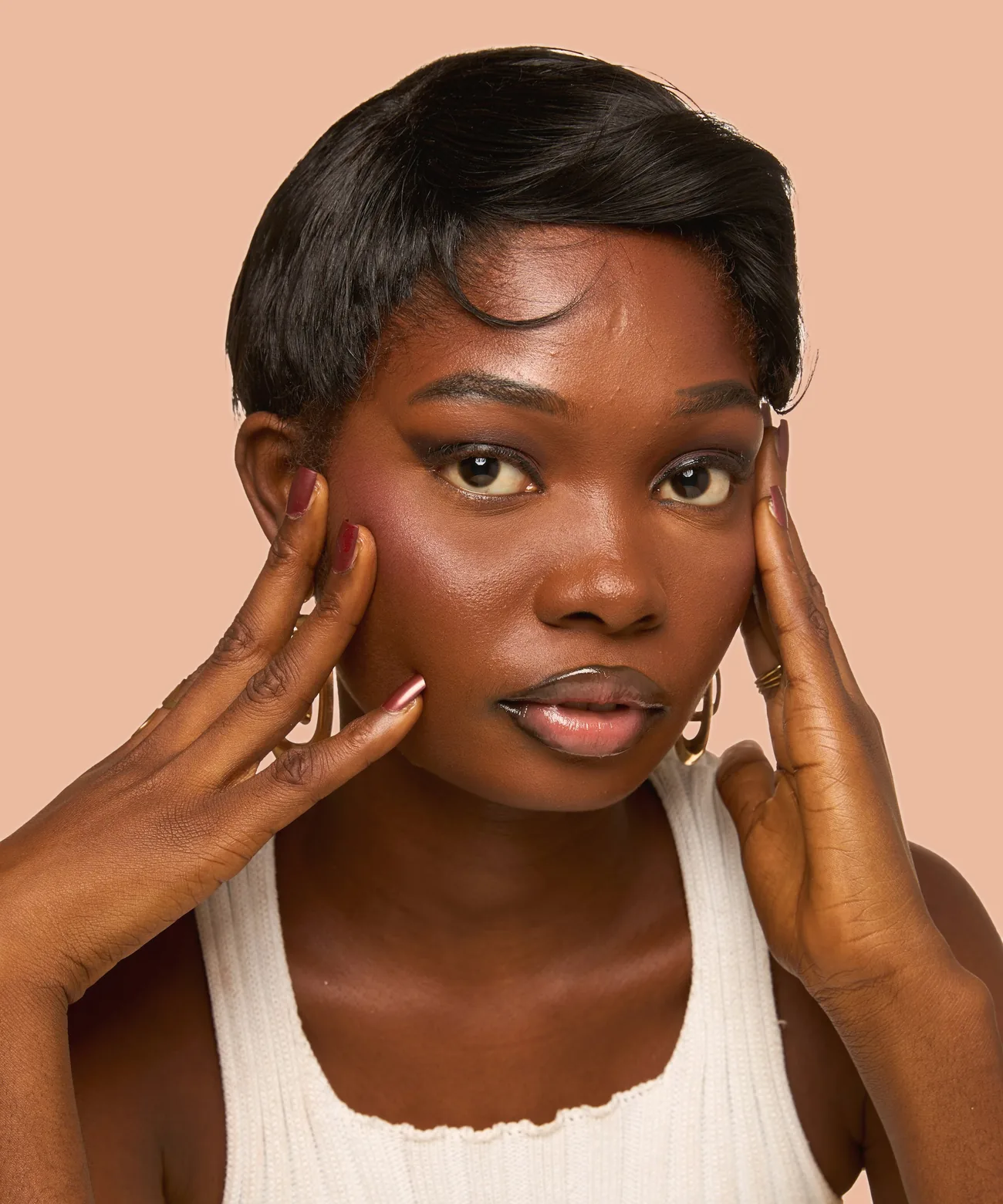 A woman with glowing skin, hands framing her face, against a peach backdrop
